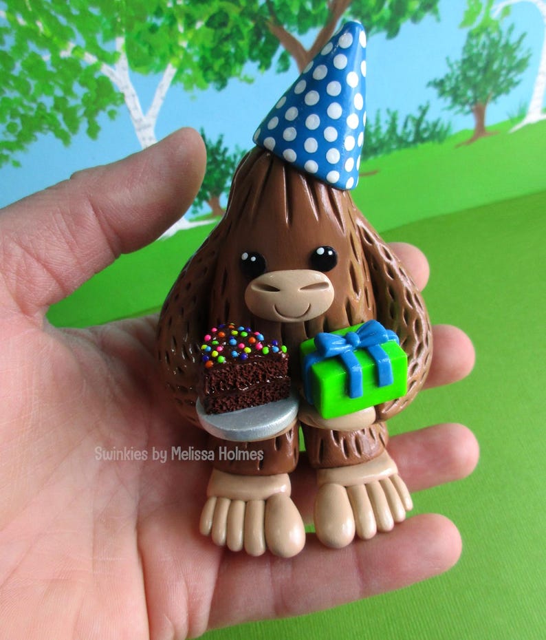 May include: A brown Bigfoot figurine wearing a blue party hat with white polka dots. The figurine holds a small chocolate cake with colorful sprinkles and a green gift box with a blue bow. The text "Swinkies by Melissa Holmes" is visible.
