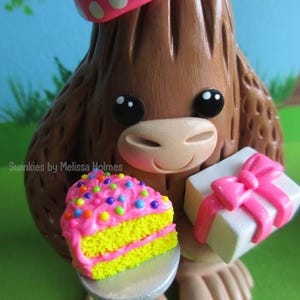 May include: A brown figurine of a cartoon character wearing a pink polka-dot party hat. The character holds a slice of cake with pink frosting and colorful sprinkles, and a white gift box with a pink bow. The text "Swinkies by Melissa Holmes" is visible.