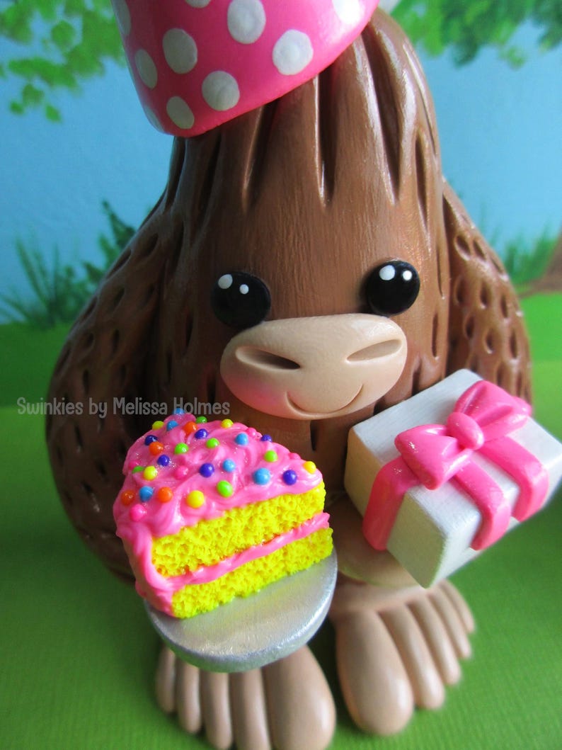 May include: A brown figurine of a cartoon character wearing a pink polka-dot party hat. The character holds a slice of cake with pink frosting and colorful sprinkles, and a white gift box with a pink bow. The text "Swinkies by Melissa Holmes" is visible.