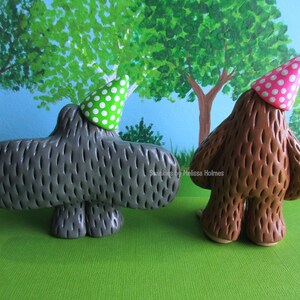 May include: Two whimsical ceramic figures, one gray and one brown, each wearing a party hat. The gray figure has a green polka dot hat, while the brown figure wears a pink polka dot hat. They stand on a green surface with a painted tree backdrop.