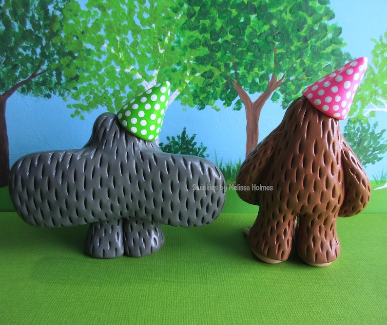 May include: Two whimsical ceramic figures, one gray and one brown, each wearing a party hat. The gray figure has a green polka dot hat, while the brown figure wears a pink polka dot hat. They stand on a green surface with a painted tree backdrop.