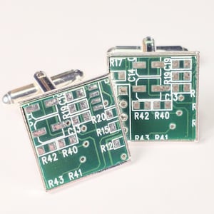 May include: A pair of silver cufflinks with a green circuit board design. The circuit board has text on it, including "R17", "C14", "R19", "C19", "C13", "R20", "R15", "R12", "R42", "R40", "R43", and "R41".