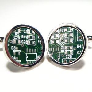 May include: A pair of silver cufflinks featuring a green circuit board design. The circuit board includes text such as "R25", "C20", "R4", "C3", "R22", "R33", "R21", "C28", "R47", "C25", "R5", "D3", and "R2".
