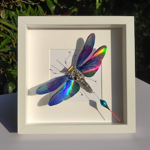 May include: A framed artwork featuring a metallic dragonfly with iridescent wings. The dragonfly is made of metal and has a detailed, intricate design. The wings are a vibrant blue, green, and purple.