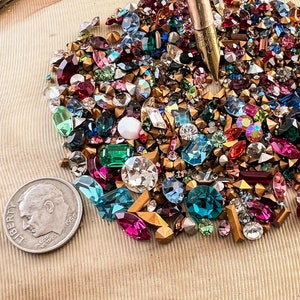 May include: A large assortment of colorful rhinestones and glass beads in various shapes and sizes, displayed on a gold-toned circular tray with a textured surface. A silver coin is included for scale.