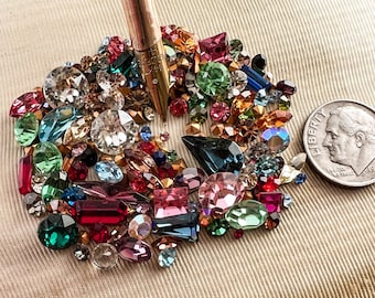Puede incluir: Una colección de pedrería de colores en varias formas, incluyendo transparente, rojo, verde, azul y rosa. Una moneda antigua está al lado de la pedrería. Un bolígrafo está en el centro de la pedrería.