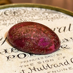 May include: A close-up of a red and gold oval-shaped cabochon with a swirling pattern. The cabochon is sitting on a vintage label with the text "All for HW Beauty" and "Prepared only by the sign C.F.  Hubert Ayer Genuine York Paris".
