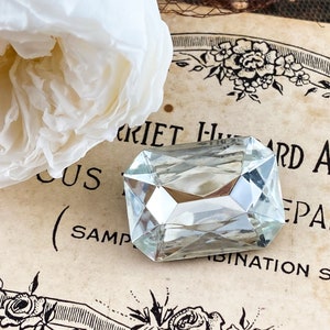 Puede incluir: Una gema de vidrio transparente con forma rectangular, descansando sobre un papel vintage con el texto "Harriet Hubbard Ayer Preparation (Sample Combination Set)".