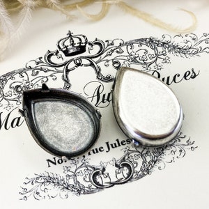 May include: Two silver teardrop-shaped locket pendants, open and closed, on a white surface with black floral designs and the text "Ma Auduces No. rue Jules".