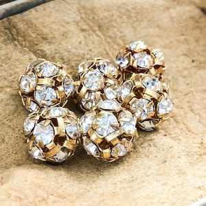 May include: Six gold-toned round beads with clear rhinestones. The beads are arranged in a cluster on a brown surface.