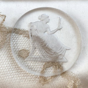 May include: A white glass cameo featuring a woman in a flowing gown holding a flower. The cameo is circular and has a raised design.