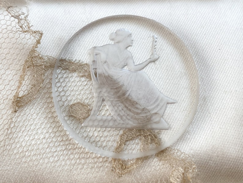 May include: A white glass cameo featuring a woman in a flowing gown holding a flower. The cameo is circular and has a raised design.