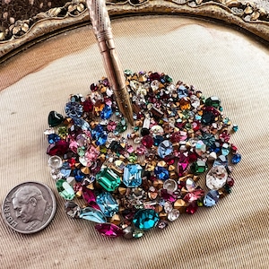 May include: A collection of colorful glass rhinestones in various shapes and sizes, including round, square, and teardrop, arranged in a circular pattern on a white fabric background.