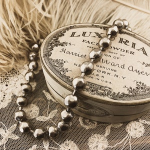 May include: A vintage round tin box with a white background and black lettering that reads "LUXURIA FACE POWDER PRED ONLY BY SIGN OF Harriet Hubbard Ayer GENUINE NEW YORK, N.Y. U.S.A. MADE IN U.S.A."  A silver beaded necklace is draped over the box.