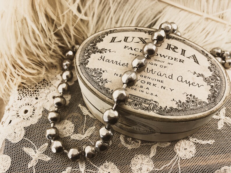 May include: A vintage round tin box with a white background and black lettering that reads "LUXURIA FACE POWDER PRED ONLY BY SIGN OF Harriet Hubbard Ayer GENUINE NEW YORK, N.Y. U.S.A. MADE IN U.S.A."  A silver beaded necklace is draped over the box.