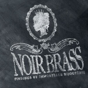 Puede incluir: Una imagen en blanco y negro de un logotipo para una joyer&iacute;a llamada "Noir Brass Findings by Immortelle Bijouterie". El logotipo presenta una imagen estilizada de la cabeza de una mujer con una corona, rodeada de un c&iacute;rculo de elementos decorativos.