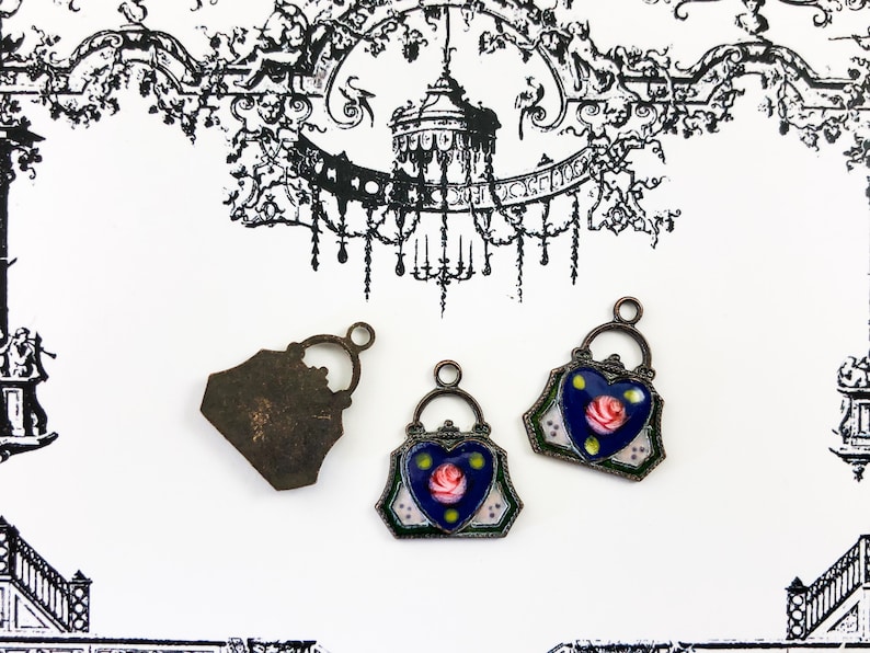 May include: Three antique-style metal charms in the shape of handbags. Two of the charms are decorated with blue enamel and a pink rose. The third charm is plain metal.
