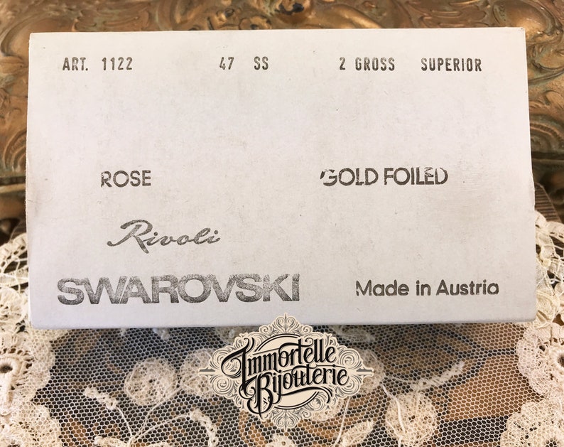 K&ouml;nnte beinhalten: Eine wei&szlig;e Schachtel mit schwarzem Text, der "ART. 1122", "47 SS", "2 GROSS SUPERIOR", "ROSE", "GOLD FOILED", "Rivoli SWAROVSKI" und "Made in Austria" lautet.