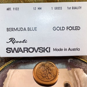 K&ouml;nnte beinhalten: Eine wei&szlig;e Schachtel mit Text, der "ART. 1122", "12 mm", "1 GROSS", "1st QUALITY", "BERMUDA BLUE", "GOLD FOILED", "Rivoli" und "SWAROVSKI Made in Austria" lautet. Ein goldfarbenes Wachssiegel im Vordergrund.