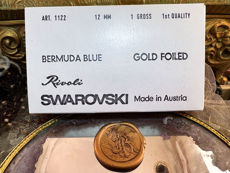 K&ouml;nnte beinhalten: Eine wei&szlig;e Schachtel mit Text, der "ART. 1122", "12 mm", "1 GROSS", "1st QUALITY", "BERMUDA BLUE", "GOLD FOILED", "Rivoli" und "SWAROVSKI Made in Austria" lautet. Ein goldfarbenes Wachssiegel im Vordergrund.