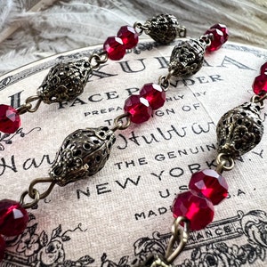 May include: Close-up of three ornate, antique-style jewelry pieces. Each piece features faceted, deep red beads and decorative, filigree metalwork. The pieces are connected by small metal chains. The background includes text from a vintage label.