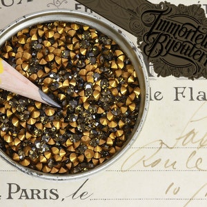 May include: A close-up of a container filled with small, gold and black rhinestones. The rhinestones are arranged in a random pattern and are all the same size. The container is made of metal and has a silver finish.