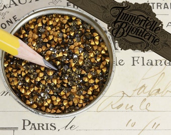 May include: A close-up of a container filled with small, gold and black rhinestones. The rhinestones are arranged in a random pattern and are all the same size. The container is made of metal and has a silver finish.