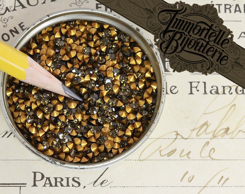 May include: A close-up of a container filled with small, gold and black rhinestones. The rhinestones are arranged in a random pattern and are all the same size. The container is made of metal and has a silver finish.