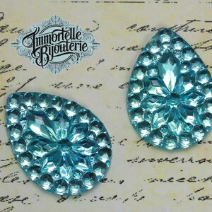 May include: Two turquoise blue teardrop-shaped glass embellishments with a raised, starburst design. The embellishments are on a white background with black writing.
