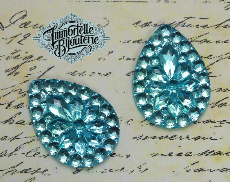 May include: Two turquoise blue teardrop-shaped glass embellishments with a raised, starburst design. The embellishments are on a white background with black writing.