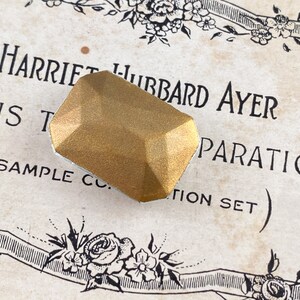 Puede incluir: Un objeto de color dorado, facetado, de forma geom&eacute;trica con una superficie ligeramente texturizada. El objeto est&aacute; sobre un trozo de papel con el texto "Harriet Hubbard Ayer Preparations (Sample Co. Edition Set)".