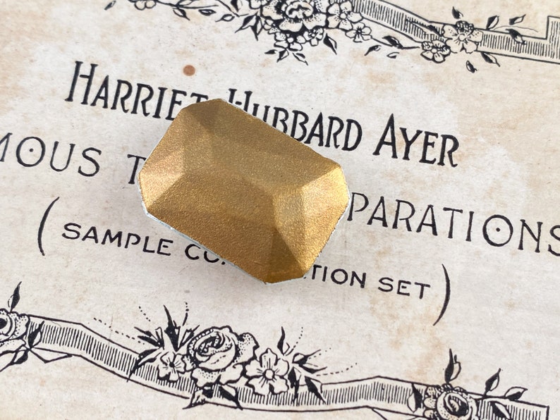 Puede incluir: Un objeto de color dorado, facetado, de forma geom&eacute;trica con una superficie ligeramente texturizada. El objeto est&aacute; sobre un trozo de papel con el texto "Harriet Hubbard Ayer Preparations (Sample Co. Edition Set)".