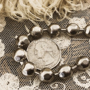 May include: A silver chain necklace with round, faceted beads. The necklace is lying on a white lace fabric with a silver US quarter coin featuring the profile of George Washington.