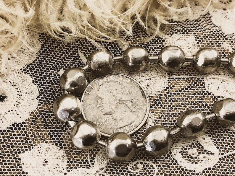 May include: A silver chain necklace with round, faceted beads. The necklace is lying on a white lace fabric with a silver US quarter coin featuring the profile of George Washington.
