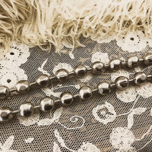 May include: Two strands of silver chain with round, faceted beads. The chain is lying on a white lace fabric with a fluffy, white feather background.