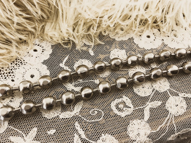 May include: Two strands of silver chain with round, faceted beads. The chain is lying on a white lace fabric with a fluffy, white feather background.