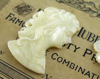 May include: A white, carved, mother-of-pearl cameo of a woman's profile with a detailed hairstyle. The cameo is a vintage style and has a pearlescent sheen.
