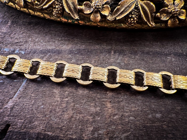 K&ouml;nnte beinhalten: Ein goldenes Kettenarmband mit geometrischem Muster. Die Kette besteht aus miteinander verbundenen ovalen Gliedern mit einem strukturierten Design.