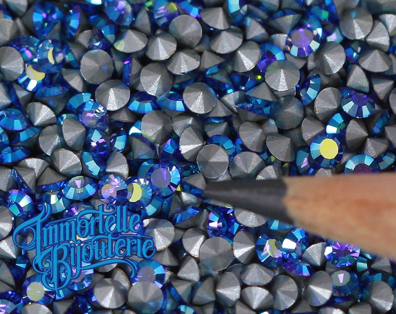 May include: A close-up image of a large quantity of small, round, iridescent blue rhinestones. The rhinestones have a silver backing and are arranged in a random pattern.