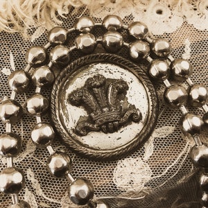 May include: A silver button with a royal crest design surrounded by a chain of silver beads. The button is on a white lace background.