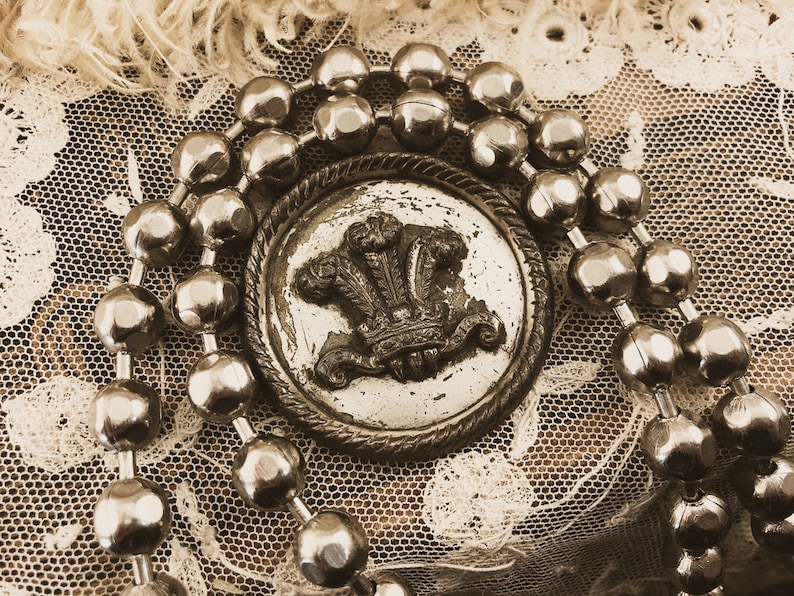 May include: A silver button with a royal crest design surrounded by a chain of silver beads. The button is on a white lace background.