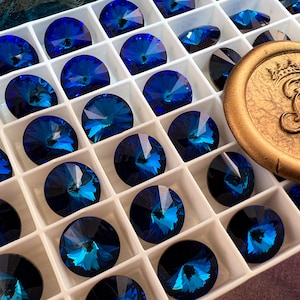 K&ouml;nnte beinhalten: Eine Anordnung von tiefblauen, facettierten Kristall-Strasssteinen in einem wei&szlig;en, unterteilten Tablett. Die kreisf&ouml;rmigen Kristalle haben einen Brillantschliff und reflektieren T&uuml;rkis- und Saphirfarben. Ein goldfarbenes Wachssiegel mit gepr&auml;gtem Design ist ebenfalls zu sehen.
