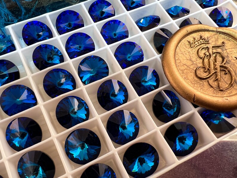 K&ouml;nnte beinhalten: Eine Anordnung von tiefblauen, facettierten Kristall-Strasssteinen in einem wei&szlig;en, unterteilten Tablett. Die kreisf&ouml;rmigen Kristalle haben einen Brillantschliff und reflektieren T&uuml;rkis- und Saphirfarben. Ein goldfarbenes Wachssiegel mit gepr&auml;gtem Design ist ebenfalls zu sehen.