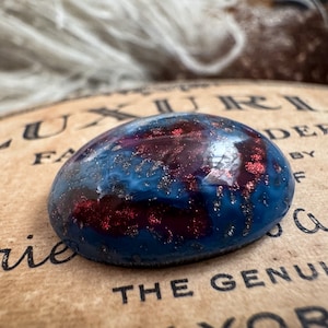 May include: A close-up of a round, blue and red resin cabochon with gold glitter. The cabochon has a galaxy-like appearance with swirls of blue, red, and gold.