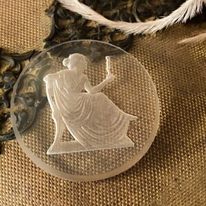 May include: A clear glass cameo featuring a woman in a flowing gown holding a candle. The cameo is set against a brown fabric background.