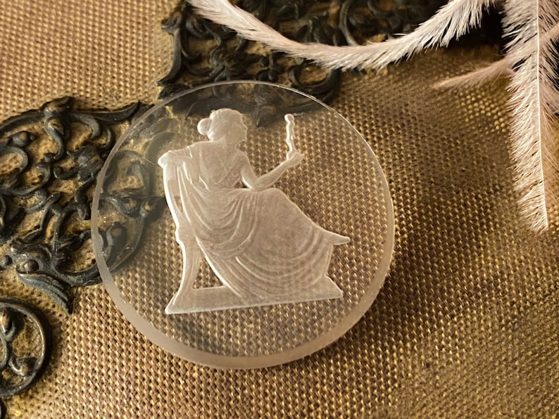 May include: A clear glass cameo featuring a woman in a flowing gown holding a candle. The cameo is set against a brown fabric background.