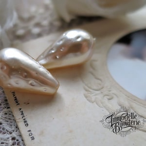 May include: Two pearlescent white teardrop-shaped beads with a textured surface. The beads are on a white surface with the text "Payment Applied For" and "Immortelle Bijouterie".