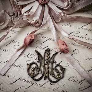 May include: A vintage-style composition featuring a brass monogram, delicate pink ribbons, and small rose accents. The background is a cream-colored paper with handwritten script, creating a romantic and antique aesthetic.