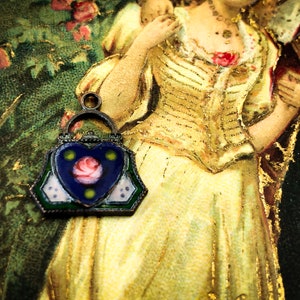 May include: A vintage enamel charm in the shape of a handbag with a heart-shaped front panel. The heart is blue with a pink rose and green accents. The charm is silver-toned with a green, white, and blue enamel design.