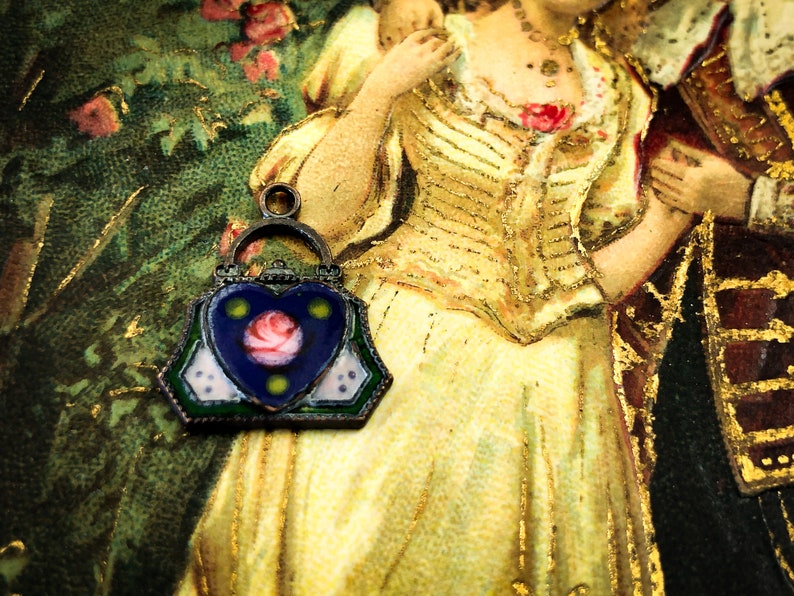 May include: A vintage enamel charm in the shape of a handbag with a heart-shaped front panel. The heart is blue with a pink rose and green accents. The charm is silver-toned with a green, white, and blue enamel design.